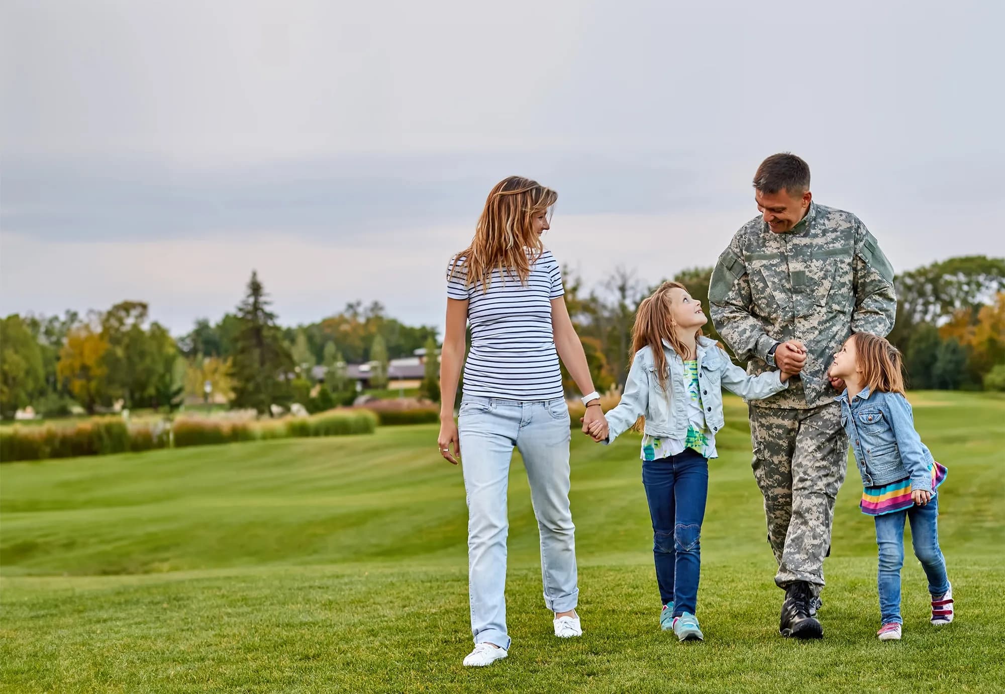 A soldier in camouflage walking across a lawn hand in hand with his two young daughters, their mother beside them
