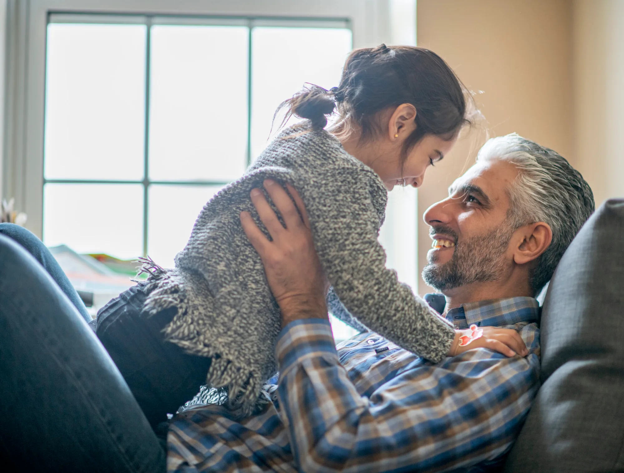 A grey-haired man lying back on a sofa, smiling up at a young girl in a knit sweater leaning over him