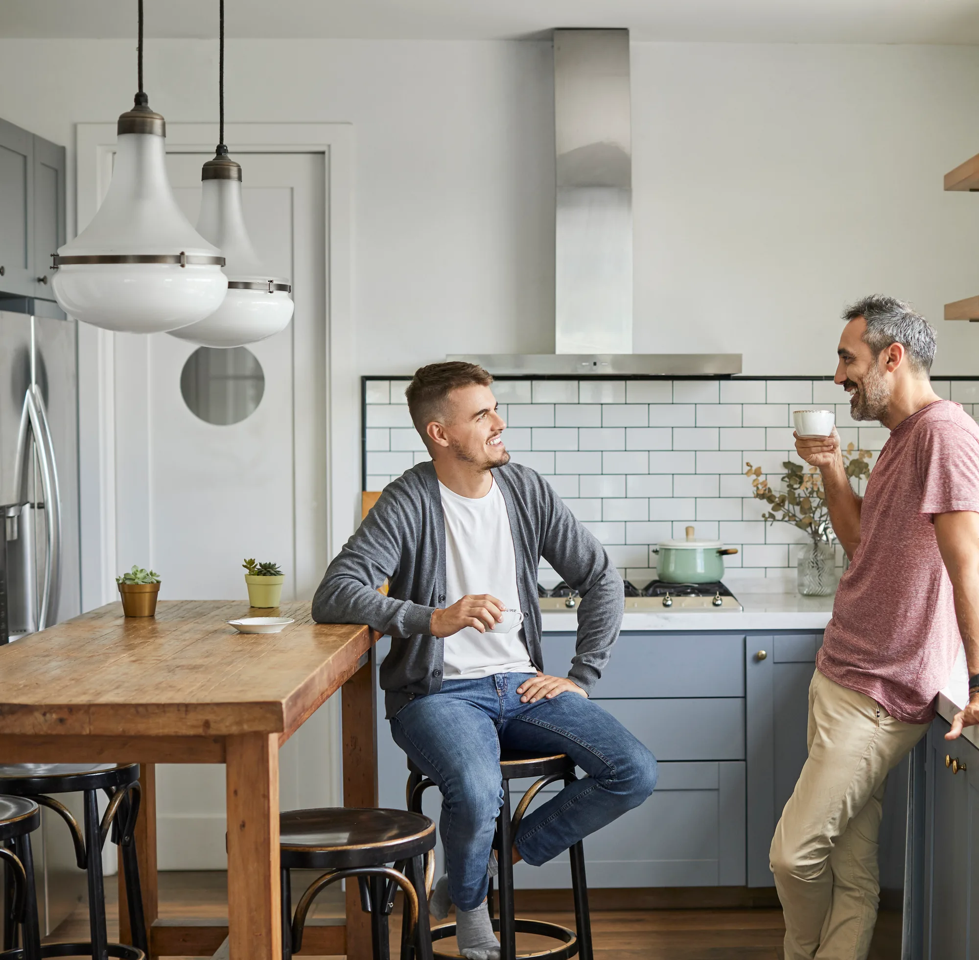 Two men talking over coffee in a sunlit kitchen, one seated at the island and the other leaning against the counter with a mug