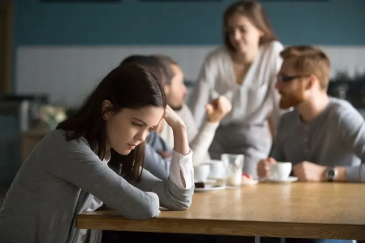 A woman looking down with her head resting on one hand while people talk nearby