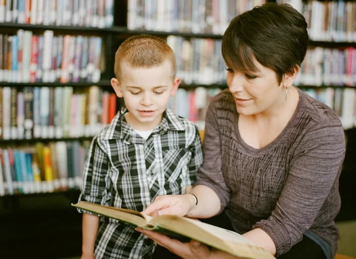 A woman and a young boy read an open book together in front of library shelves