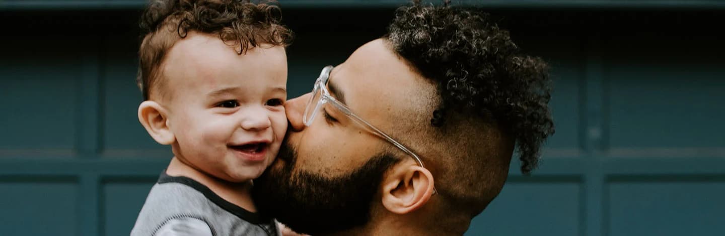 A father wearing glasses kisses his smiling young son on the cheek