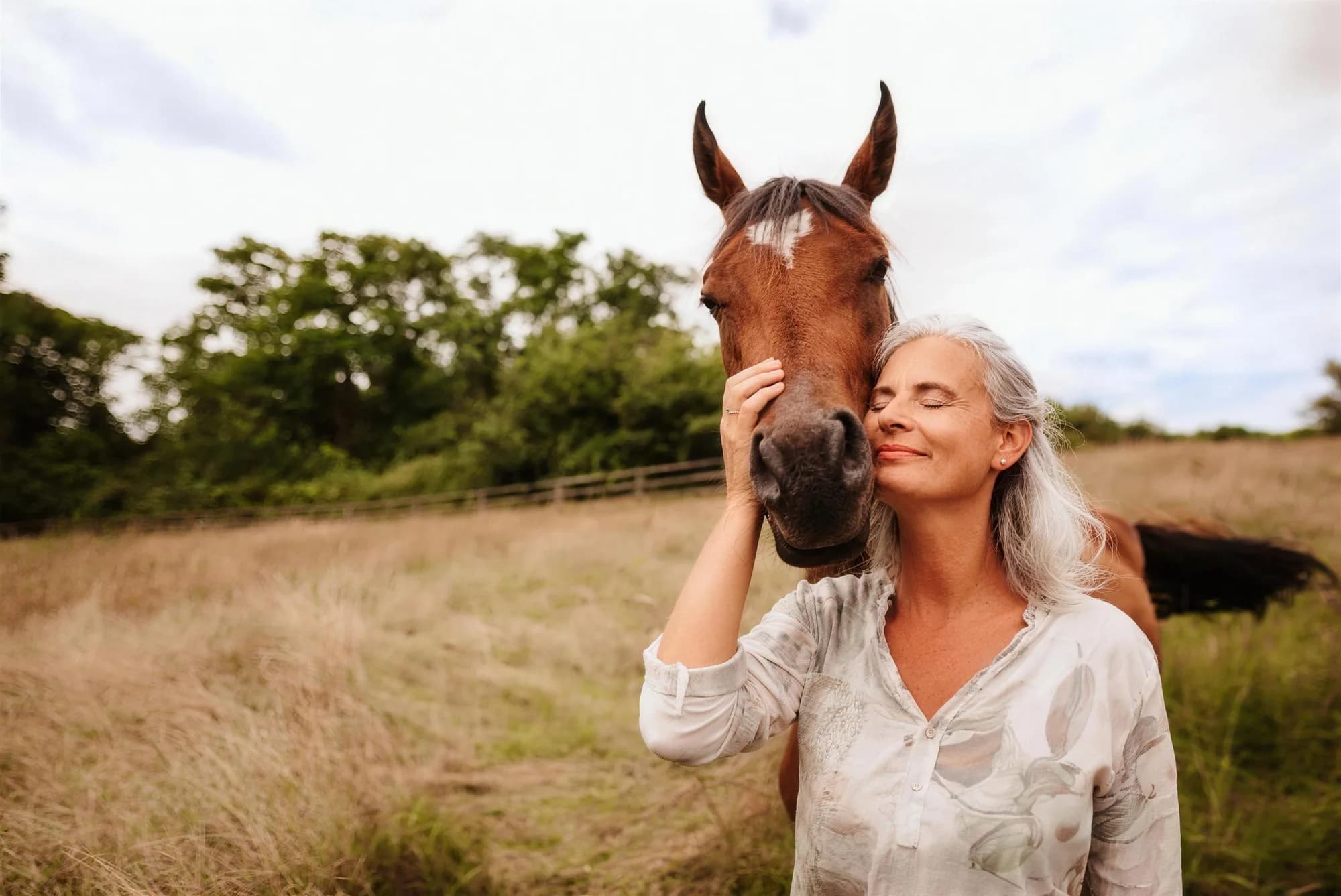 A silver-haired woman in a summer meadow, eyes closed and smiling, resting her cheek against a horse's muzzle